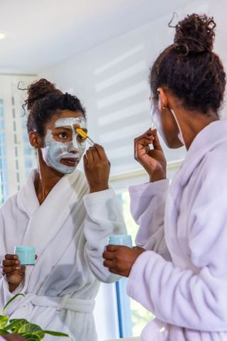 African american woman applying facial mask with brush at vanity mirror in white bathrobe