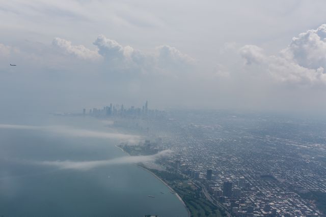 Foggy aerial view of a large city skyline with lakefront and urban sprawl. Skyscrapers partially obscured by fog create mysterious and tranquil atmosphere. Useful for content related to urban life, weather patterns, travel, tourism, and city environments.