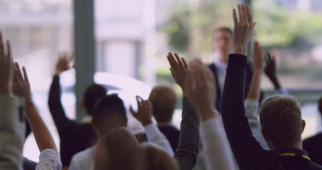 People in an audience express interest and readiness to engage by raising their hands during a seminar or workshop. This is suitable for portraying active participation, engagement, and collaboration in educational or business settings. It can be used for content related to learning environments, corporate training, team-building activities, and interactive conferences.