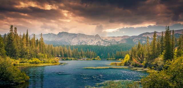 This image depicts a beautiful scenic landscape featuring a calm lake surrounded by trees, with mountains in the background under a dramatic sunset sky. Ideal for nature enthusiasts, travel blogs, outdoor adventure promotions, and environmental conservation awareness. Perfect for desktop wallpapers, digital prints, and calendar covers.