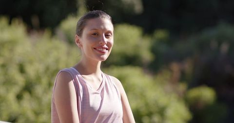 Sunlit young woman smiling and posing outdoors in light pink tank top with bokeh background