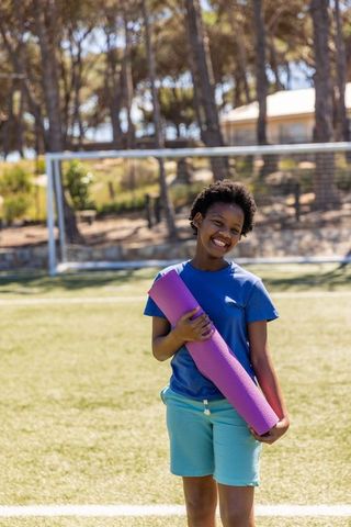 African american teen smiling and holding purple yoga mat on school soccer field outdoors