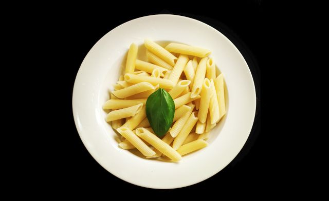 Close-up of plain penne pasta served in a white plate, garnished with a single basil leaf. Ideal for illustrating minimalist Italian cuisine, clean eating, or meal presentation concepts for restaurant menus, food blogs, or recipe websites.