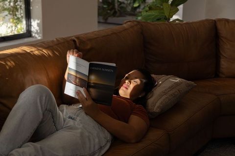 Middle-aged asian woman reclining and reading endless sunsets on leather sectional couch in sunlit l