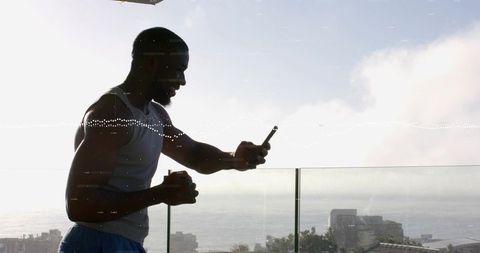 Silhouetted man checking phone with coffee cup on glass balcony overlooking ocean cityscape