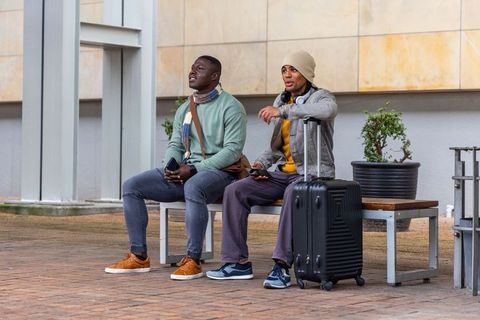 Diverse friends waiting at urban bench with wheeled suitcase and smartphones