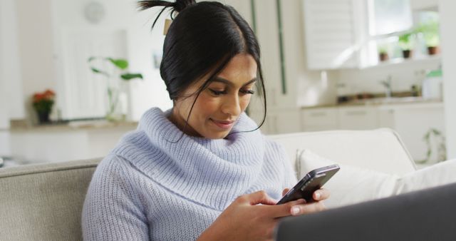 Woman in a light blue sweater using smartphone while sitting on a comfortable sofa at home. Perfect for themes related to modern lifestyle, technology use, home comfort, relaxation, and indoor leisure activities.