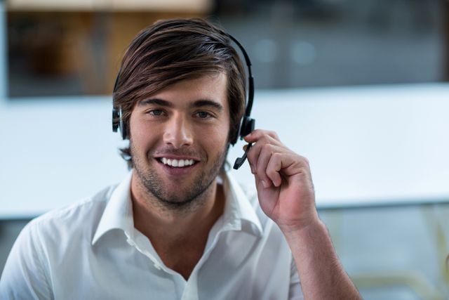 Young male customer service representative wearing headset, smiling at camera, in modern office environment. Ideal for use in business, customer support, and telecommunication service promotions. Perfect for illustrating concepts of customer service, help desk, and professional communication.