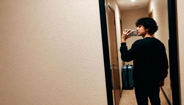Teen taking mirror selfie in hallway with teal suitcase and smartphone