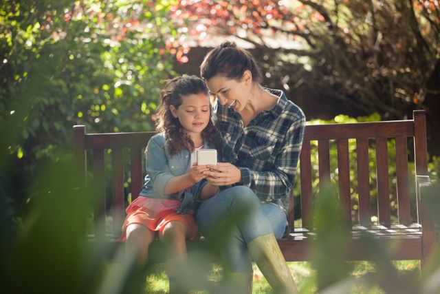 Girl showing mobile phone to mother while sitting on wooden bench at backyard