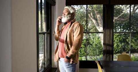 Senior Man with White Beard Talking on Phone by Sunny Window at Home
