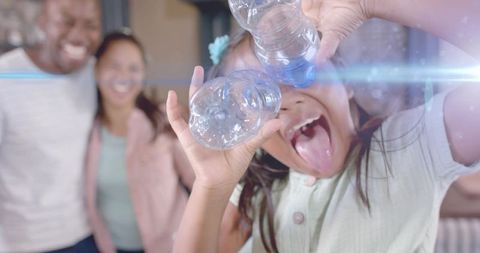 Playful child using plastic bottles as binoculars while parents laugh in cozy kitchen