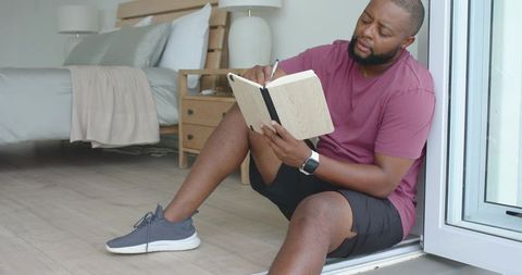African american man sitting on bedroom floor writing in notebook at open doorway journaling