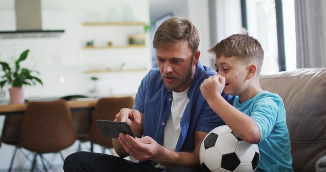 Father and son sitting on couch in living room, focusing on smartphone. Boy holding soccer ball, showing interest in video. Scene can be used for family, digital technology, or home lifestyle concepts.