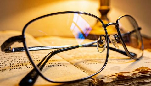 Metal-framed reading glasses resting on aged open book with warm lamp reflection