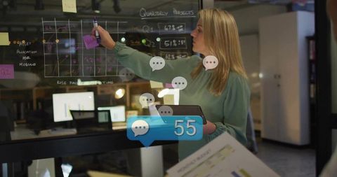 Female leader drawing strategy on glass whiteboard, holding tablet in modern office