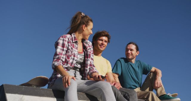 A group of three friends is sitting on steps with a bright blue sky in the background. They are casually dressed in jeans and T-shirts, engaging in conversation and smiling at each other. One of them is holding a skateboard, suggesting a youth and active lifestyle. This image can be used in contexts related to friendship, social gatherings, outdoor activities, or youth lifestyle advertisements.