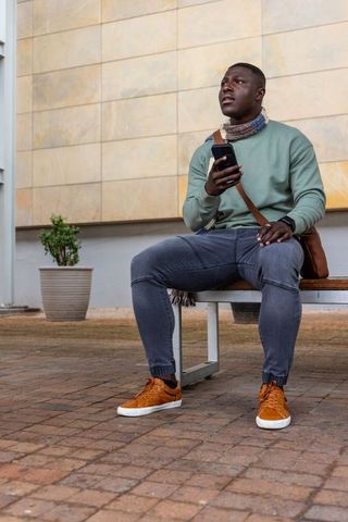 African american man sitting on bench holding smartphone and satchel in urban courtyard