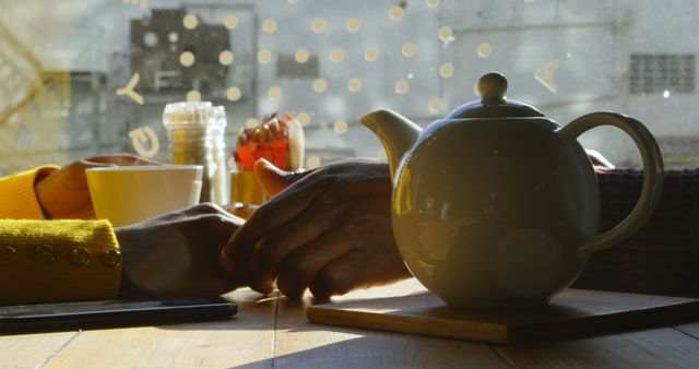 A cozy scene features a teapot on a wooden table with two people, one Caucasian and one African American, holding hands in the background, with copy space. Warm sunlight filters through, enhancing the intimate and comfortable atmosphere of a shared tea moment.