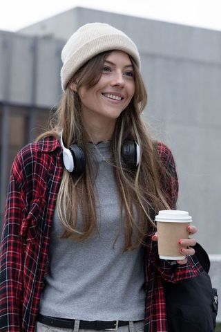 Smiling urban woman holding coffee wearing beanie and headphones in casual streetwear