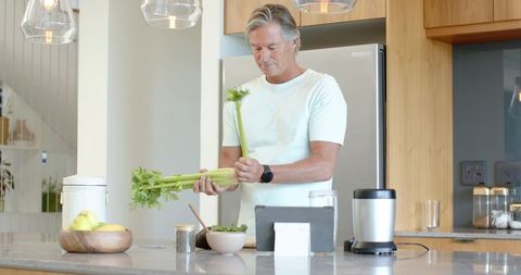 Senior man preparing celery on kitchen island using tablet in modern minimalist kitchen