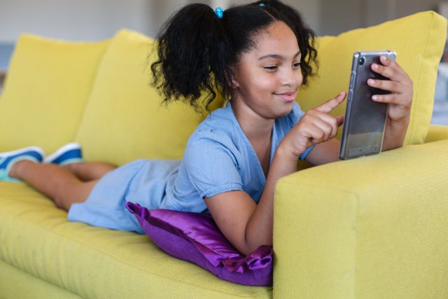 Young girl lying on a yellow couch, using a digital tablet. Ideal for educational content, technology use in children, or relaxation themes. Suitable for articles, blogs, or advertisements focusing on childhood learning, digital education, or comfortable home environments.