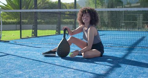 Smiling african american woman sitting on blue padel court holding paddle and ball