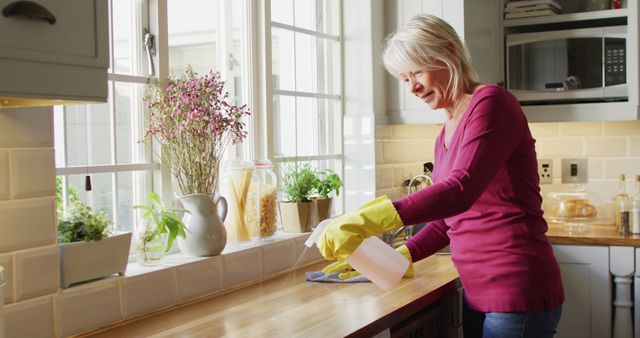 Older woman, wearing pink sweater and yellow gloves, smiling while cleaning kitchen counter with a spray bottle. Bright kitchen with sunlight streaming through windows and houseplants on windowsill. Ideal for content related to housekeeping, senior lifestyle, home hygiene, and domestic chores.