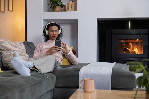 Nonbinary person relaxing on sofa with headphones and smartphone by cozy fireplace