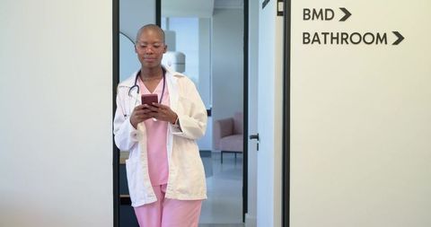 African clinician standing in doorway wearing pink scrubs and lab coat using smartphone