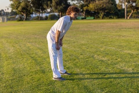 Cricket player leaning forward hands on knees catching breath on sunlit green sports field