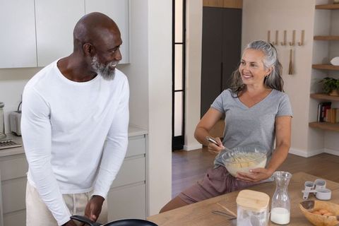 Smiling mature couple cooking together in kitchen, whisking batter and holding frying pan