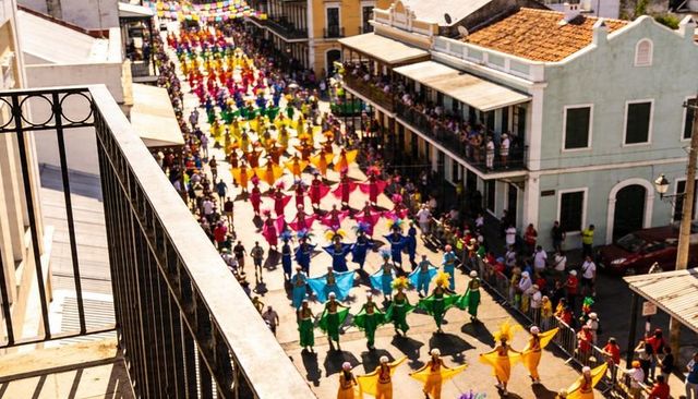 Dancers marching in rainbow capes down narrow colonial street, balcony viewpoint showing crowd