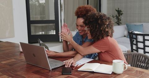 Diverse female friends celebrating success on laptop while collaborating from home
