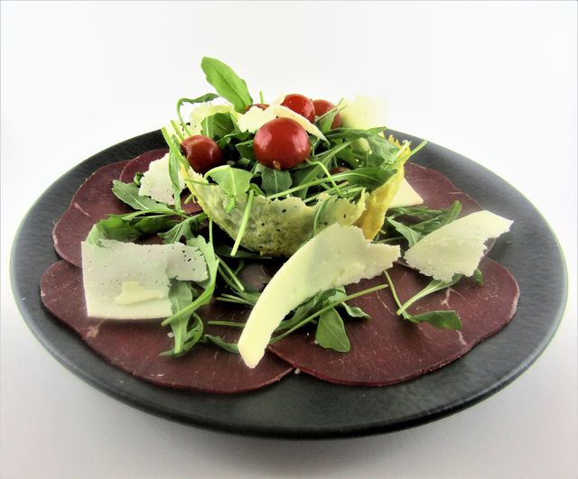 A beautifully plated antipasto featuring a bed of thinly sliced cured meat topped with fresh arugula, cherry tomatoes, and slices of parmesan cheese served on a black plate. Perfect for culinary blogs, restaurant menus, healthy eating promotions, or gourmet food presentations.