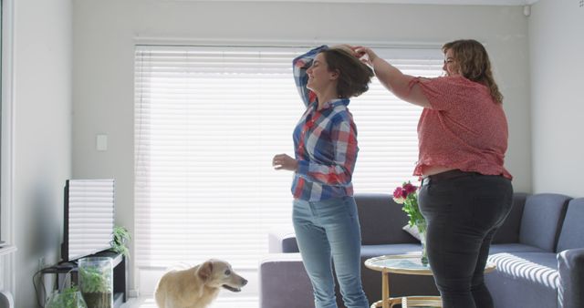 Two women engaged in a casual moment fixing hair in a bright, modern living room with a pet dog nearby. Ideal for uses related to friendship, daily life, lifestyle blogs, or pet-related content.