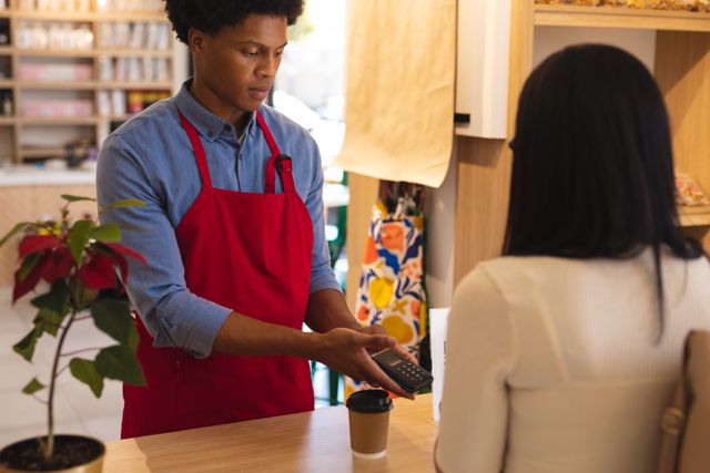 Young African American male owner in red apron accepting payment from biracial female customer at a modern cafeteria checkout counter. Ideal for illustrating small business operations, customer service, retail transactions, and modern payment technology in cafes or coffee shops.
