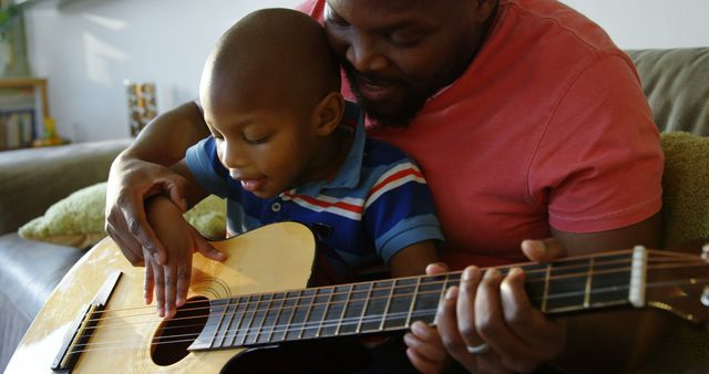 Front view of African american father teaching his son how to play guitar at home. They are sitting on the sofa. 4k