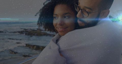 Embracing couple snuggling on seaside railing wrapped in blanket at dusk with lens flare