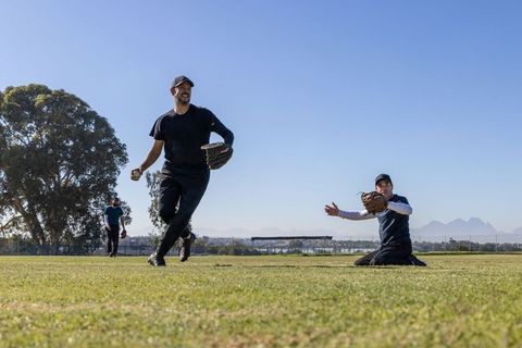 Diverse male teammates playing baseball on sunny grass field running and catching ball