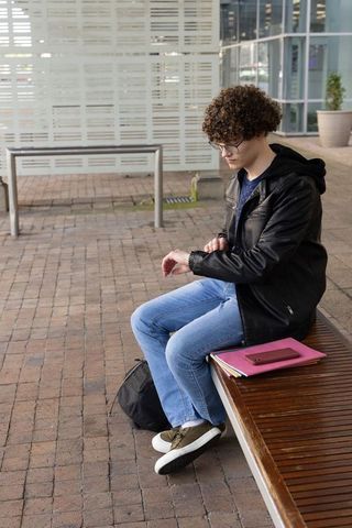 Teen male sitting on plaza bench checking wristwatch, waiting with backpack and notebooks