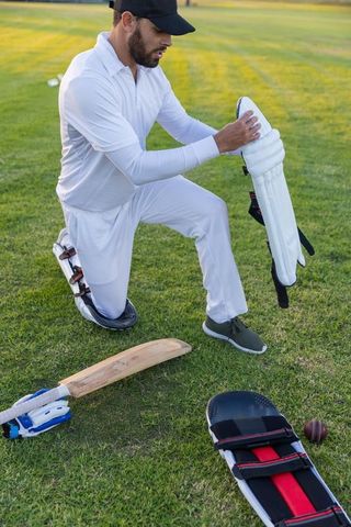 Kneeling cricketer adjusting leg pads on sunlit cricket field with bat gloves and ball