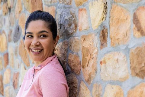 Smiling woman leaning on stone wall wearing pink active top in bright daylight