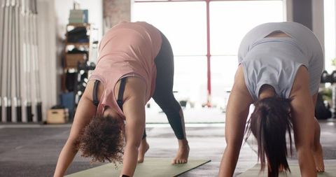 Two women performing downward-facing dog yoga together on mats in bright gym studio