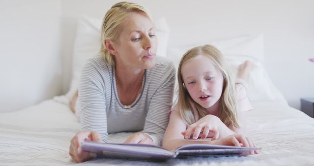 Happy caucasian mother and daughter lying on bed reading story book together. Motherhood, childhood, care and togetherness.