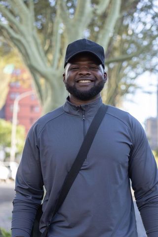 Confident african american man smiling in tree-lined urban street wearing cap and gray zip-up