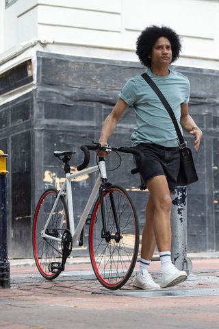 African-american commuter walking bike with red rims wearing crossbody bag on sidewalk
