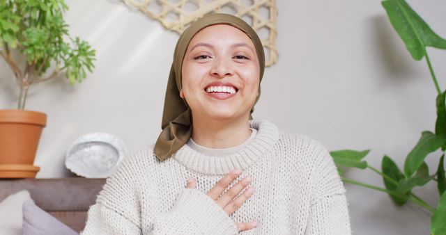 Image of happy biracial woman in hijab making image call waving to camera at home. Happiness, relaxation, communication, inclusivity and domestic life.