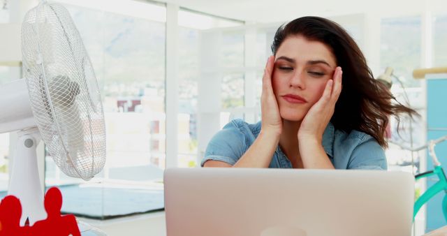 Woman sitting at desk with laptop feeling hot and exhausted while fan is blowing. Perfect for depicting office heat issues, summer work stress, or cooling products.