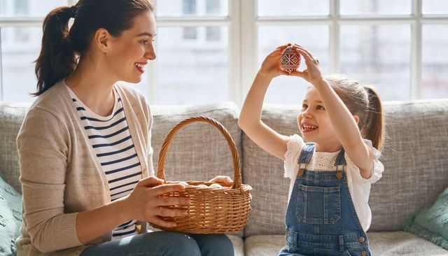 Smiling mother and daughter decorating ornate easter egg together on cozy sunlit sofa